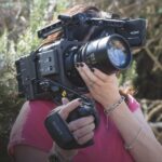 A woman wearing pink holding a large Sony Burano camera over her face. She stands outside