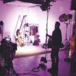 A woman sitting on a photography set surrounded by clothes. The lighting is purple