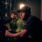 A man wearing a hat and headphones sitting in a dim lighting filming a movie