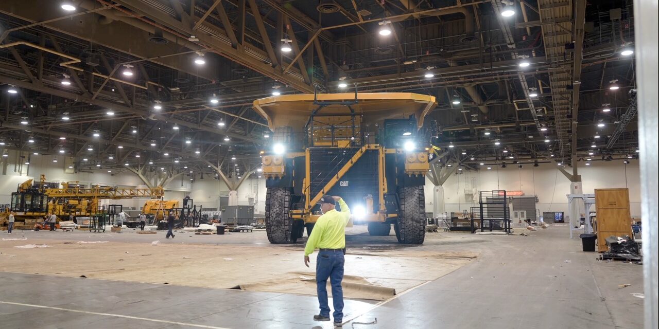 A person in a hi-vis beckons a large construction vehicle forward in a warehouse