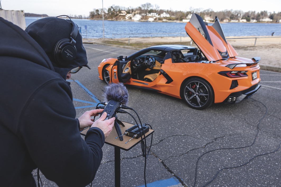 An orange sports car with wires attached to the back. The car's doors and boot are open. A man stands in the foreground with headphones and a microphone on a small table