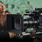 A large cinema camera overlaying a picture of a young woman in a dress looking up and smiling in a green room