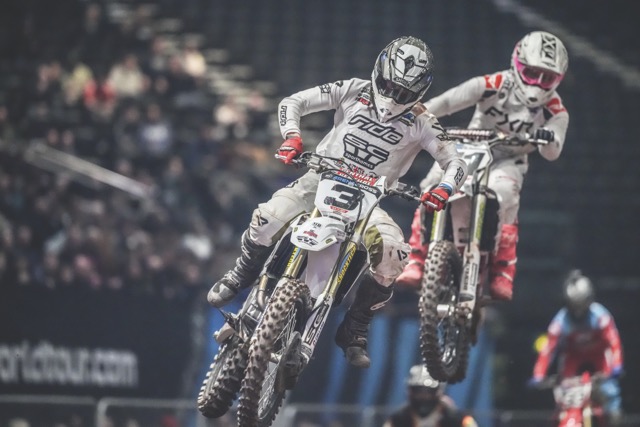 Two professional motorcyclists performing a jump in a packed arena
