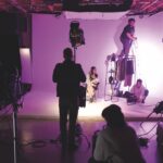 A photography set with purple lighting. A woman sits on a chair next to a clothes rack while a man stands on a ladder and a woman adjusts the set next to her