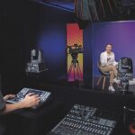 A woman sits in a film studio presenting, while a man controls aspects of the shoot from a remote control