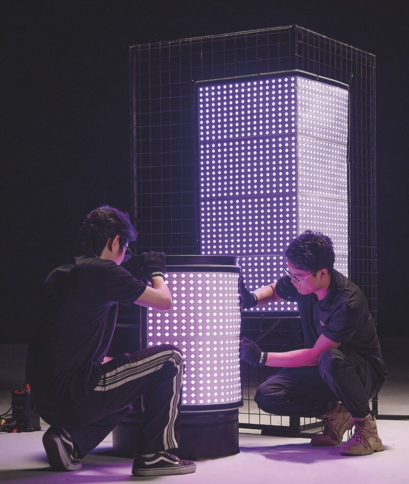 Two men wearing black gloves install a curved purple LED panel on a stage while another square panel sits behind them