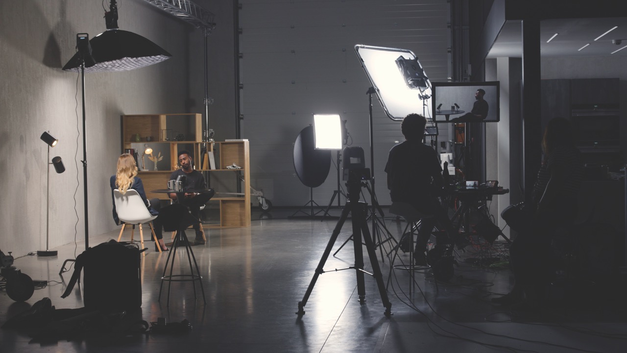 A dark filming studio with professional cameras and lights set up. Someone sits to the side of the room