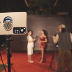 A red carpet event where women in formal dresses stand in a blurred background. A lighting device is set up on a tripod in the foreground while a man takes a photo