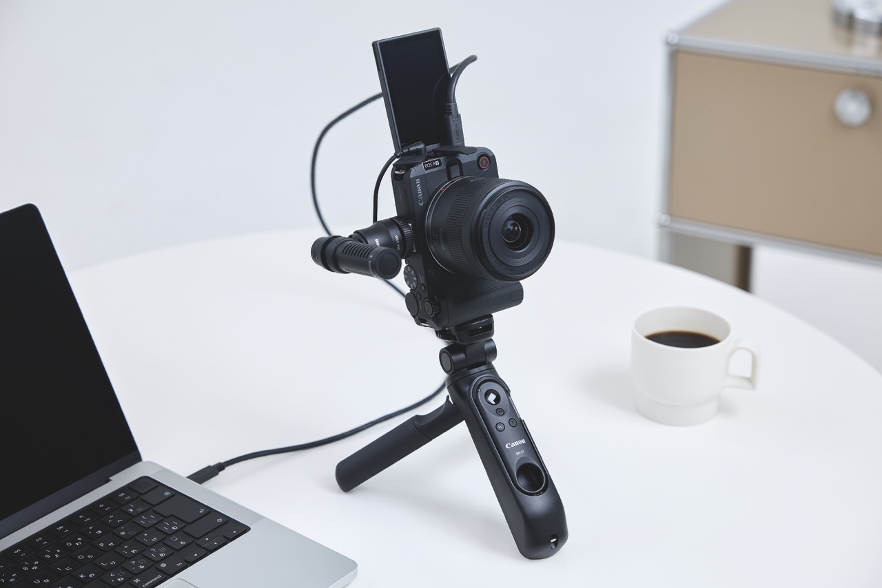 A small camera attached to a desk stand and plugged into a laptop. A coffee sits next to it on the table