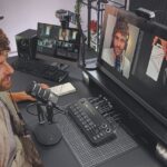 A man sitting at a desk looking at a monitor with a recording of himself on. There is a microphone and mixing desk in front of him