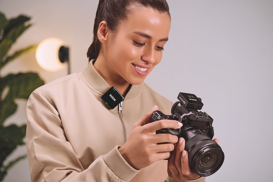 A woman holds a camera while smiling. She wears a small square microphone on her jumper