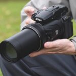 A person kneeling on the grass holding a large Nikon camera