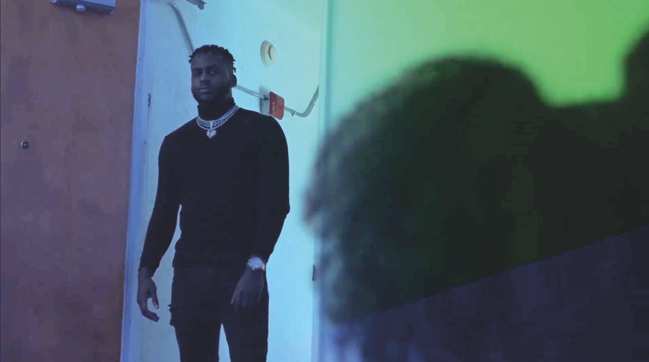 A man stands in a dim blue-green room looking towards a blurred figure in the foreground