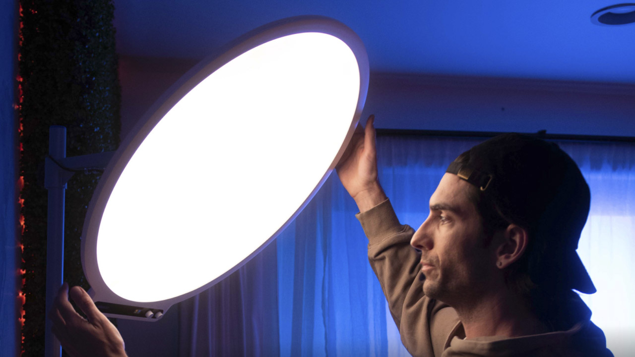 A man with a backwards cap adjusting a large circular light in a blue room
