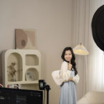 A young girl in a blue dress and white cardigan holding a white handbag. She stands in a living area between two circular lights