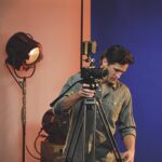 A man in a green shirt adjusting a camera on a tripod in an orange and blue room