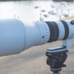 A large white lens attached to a small camera on a mount. The camera is placed on a path next to a river with birds in