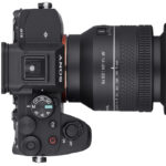 A top view of a black camera with a large lens. There are many buttons and dials on the camera