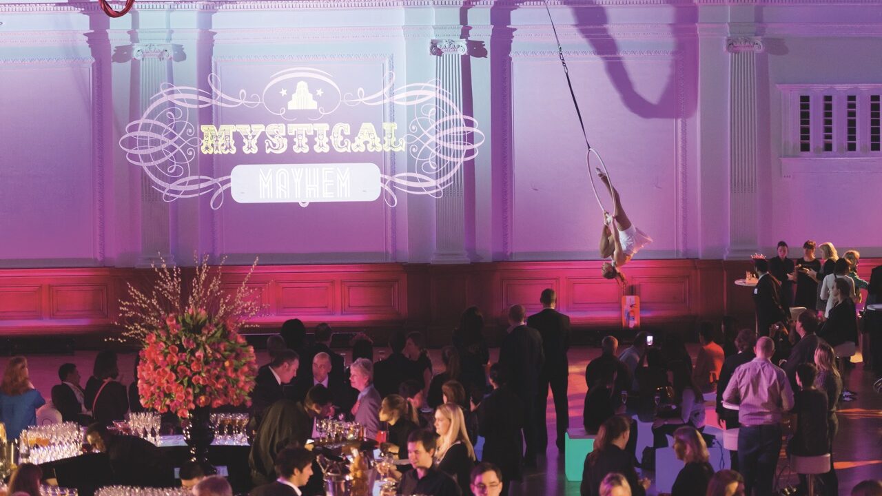 An acrobat flying over a crowd seated at round tables on a rope and hoop in a purple room