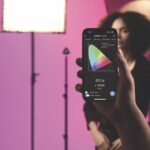 A hand holds a phone should a graph with different colours on. In the background a model poses in front of a pink background, surrounded by lights