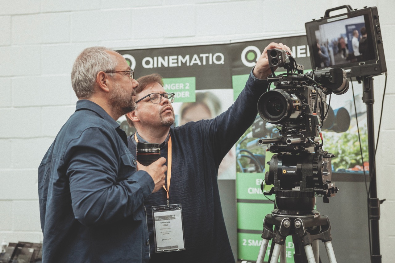 Two men adjust a camera with a large monitor next to it in a conference hall