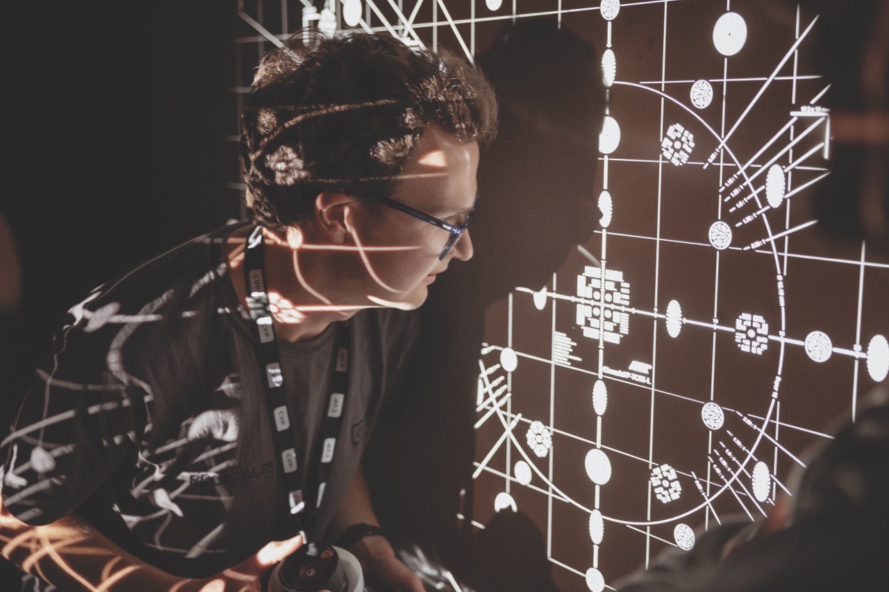 A man sits close to a wall while a projection of a lens shines on him and the wall