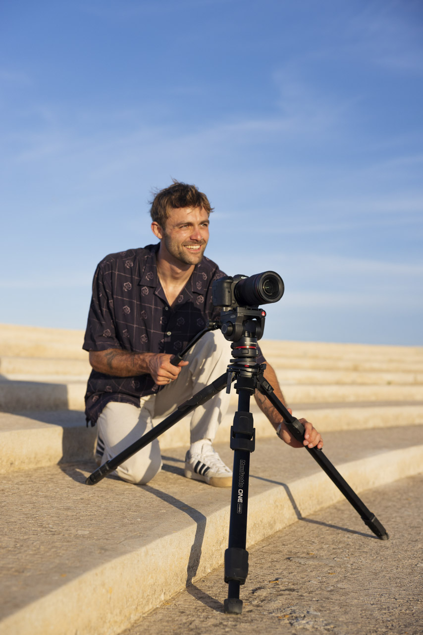 A man kneels on sandy steps against a blue background smiling. He adjusts a short tripod with a camera attached in front of him