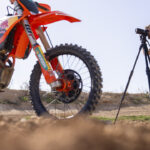 A man in the background looks through a camera lens, while shooting a red motorcycle in the foreground. The camera is placed on a tall tripod