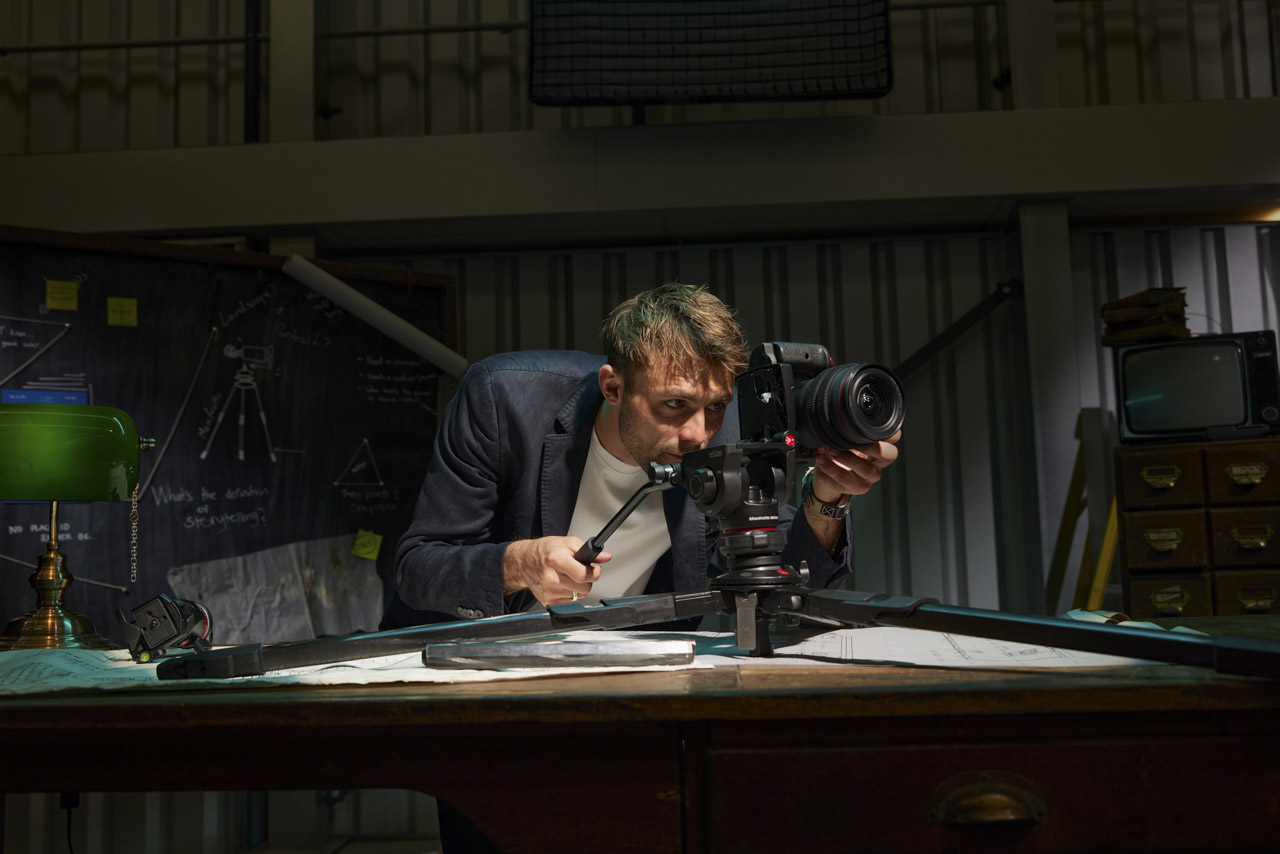 A man looks through a camera attached to a short tripod set up on a table in a dark room