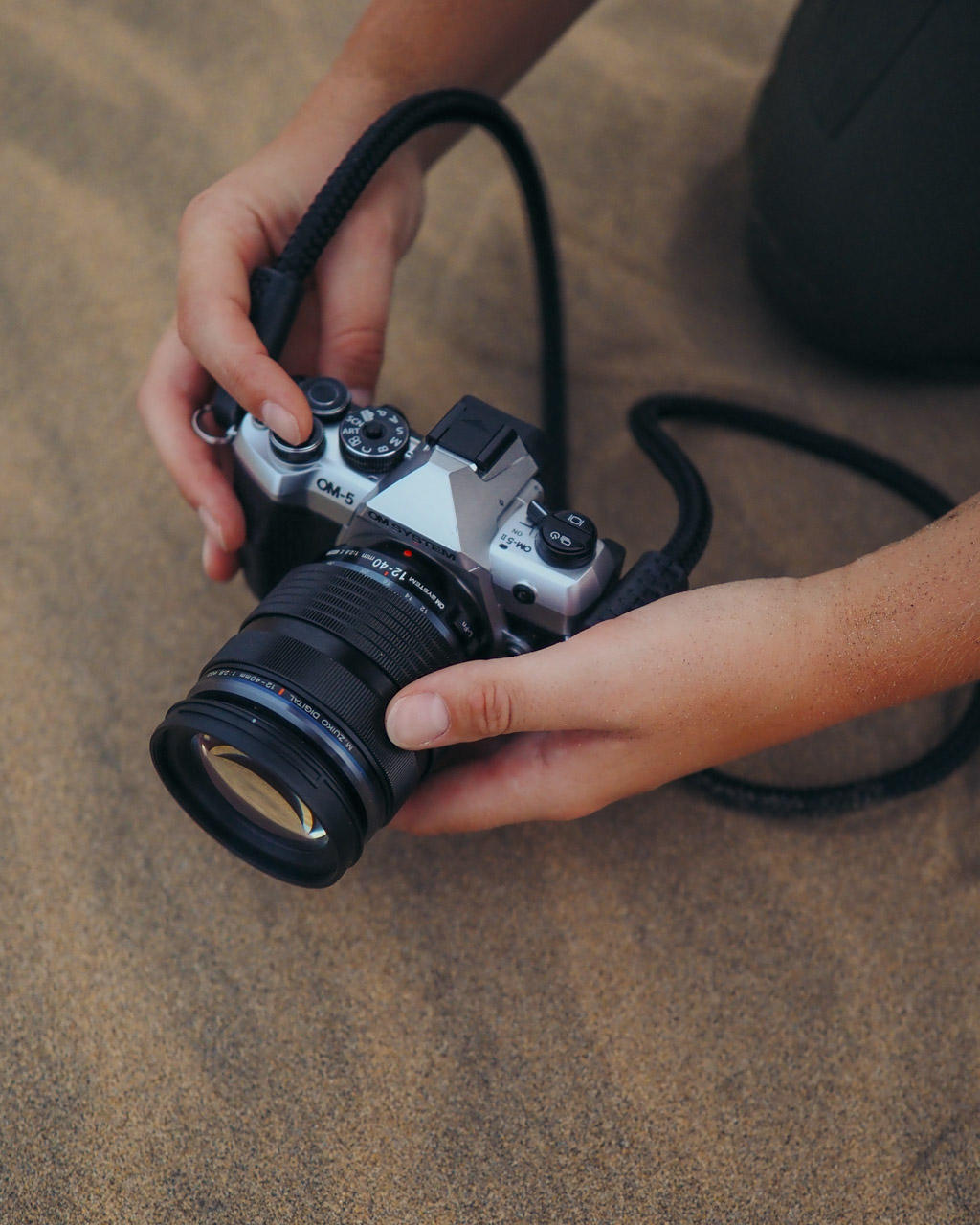 A pair of hands holds a silver and black camera close to the sandy floor. The camera has a neck strap attached