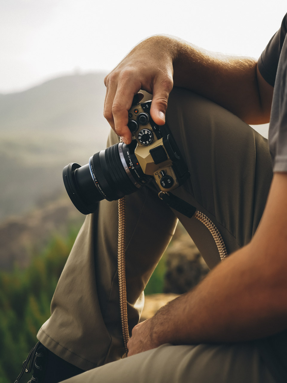 A person rests their arm on their leg while holding a camera in their hands. They sit on the edge of a mountain