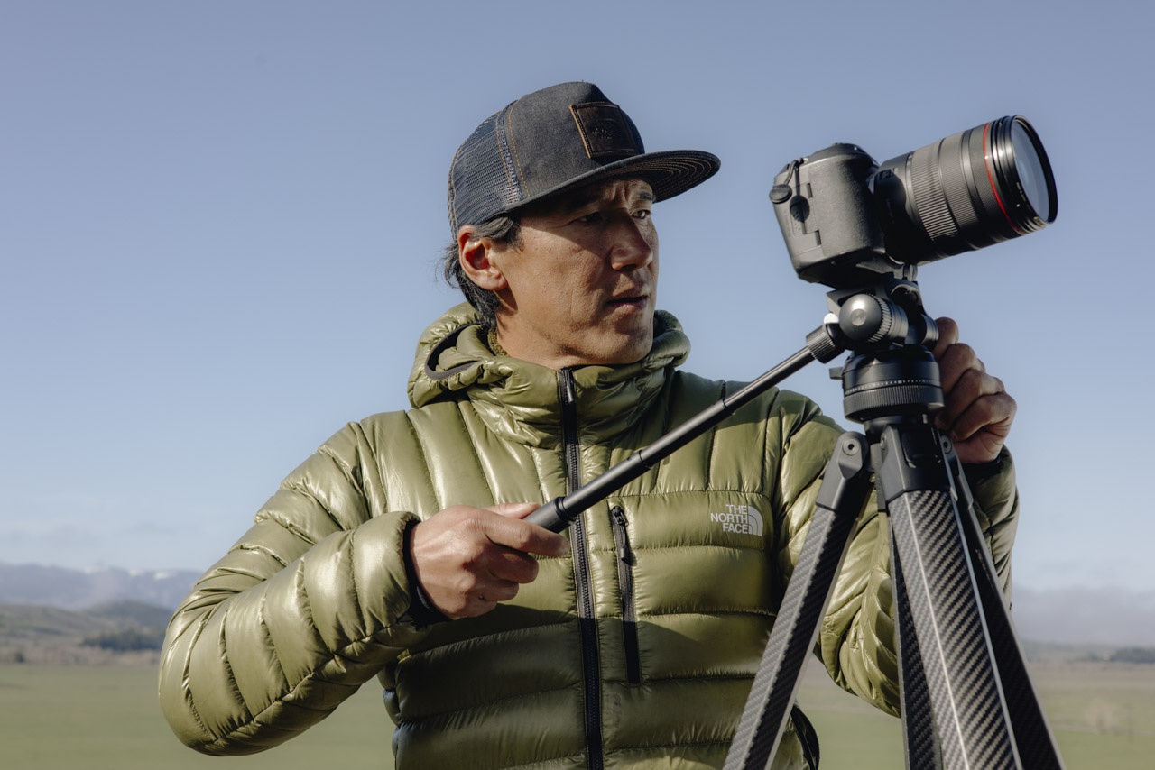 A man wearing a green coat and a cap adjusts a camera attached to a tripod outside