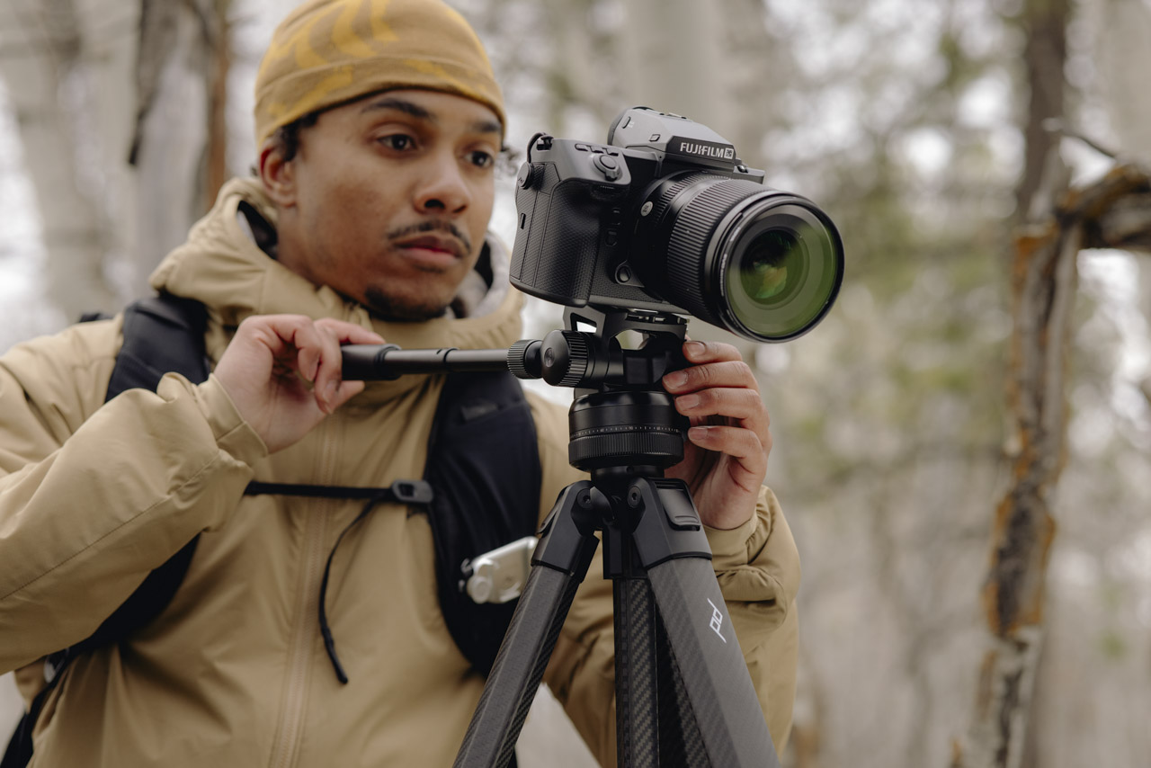 A person wearing a coat and yellow hat adjusts a camera on a tripod outside in a forest