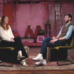 Two people sit in armchairs facing each other in a purple-lit room