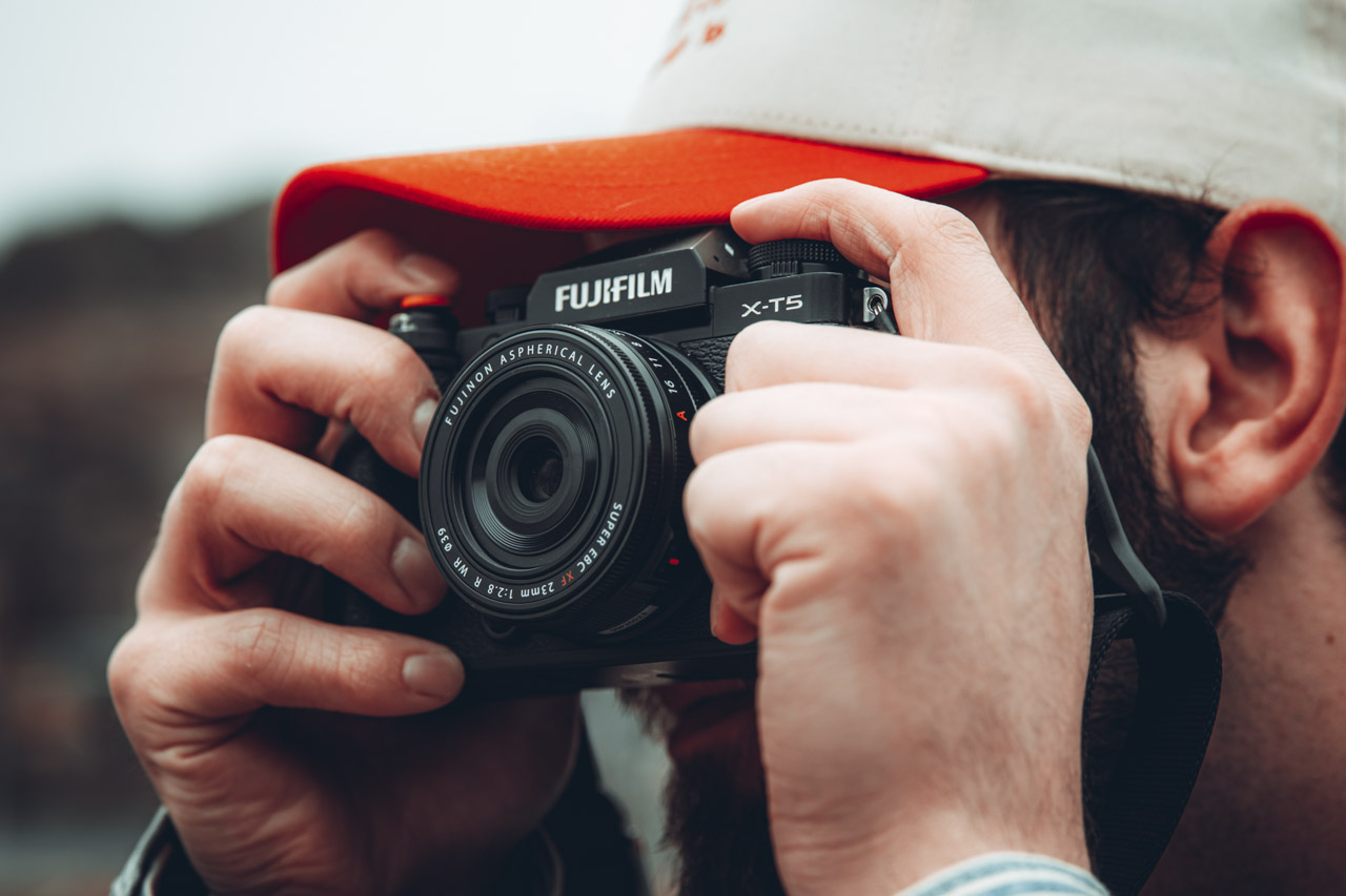 A person wearing an orange and white cap, covering their face with a small Fujifilm camera