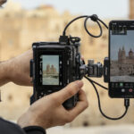 A man holding a camera vertically with a smartphone attached. He records an ornate building in front of him