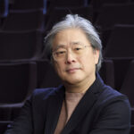 A man with glasses and grey hair wearing a dark blue blazer and sitting in a dark room
