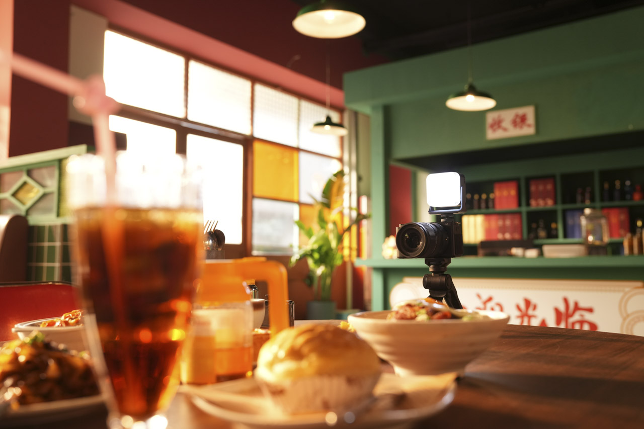 The inside of a restaurant with food and drinks in the blurred foreground and a camera set up in the background near a green counter