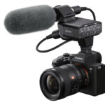 A black camera with a large flash and microphone attached to the top, against a white background