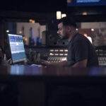 A man working at a computer next to a mixing desk in a dark room