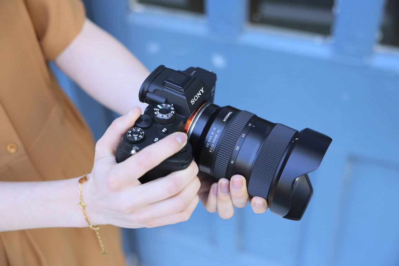 A black lens attached to a camera. It is held by a person wearing a brown dress, standing in front of a blue door