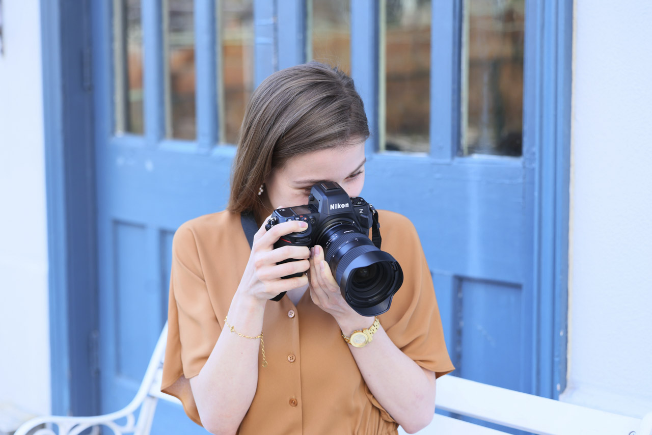 A woman wearing a brown dress looks through a camera in front of a blue door