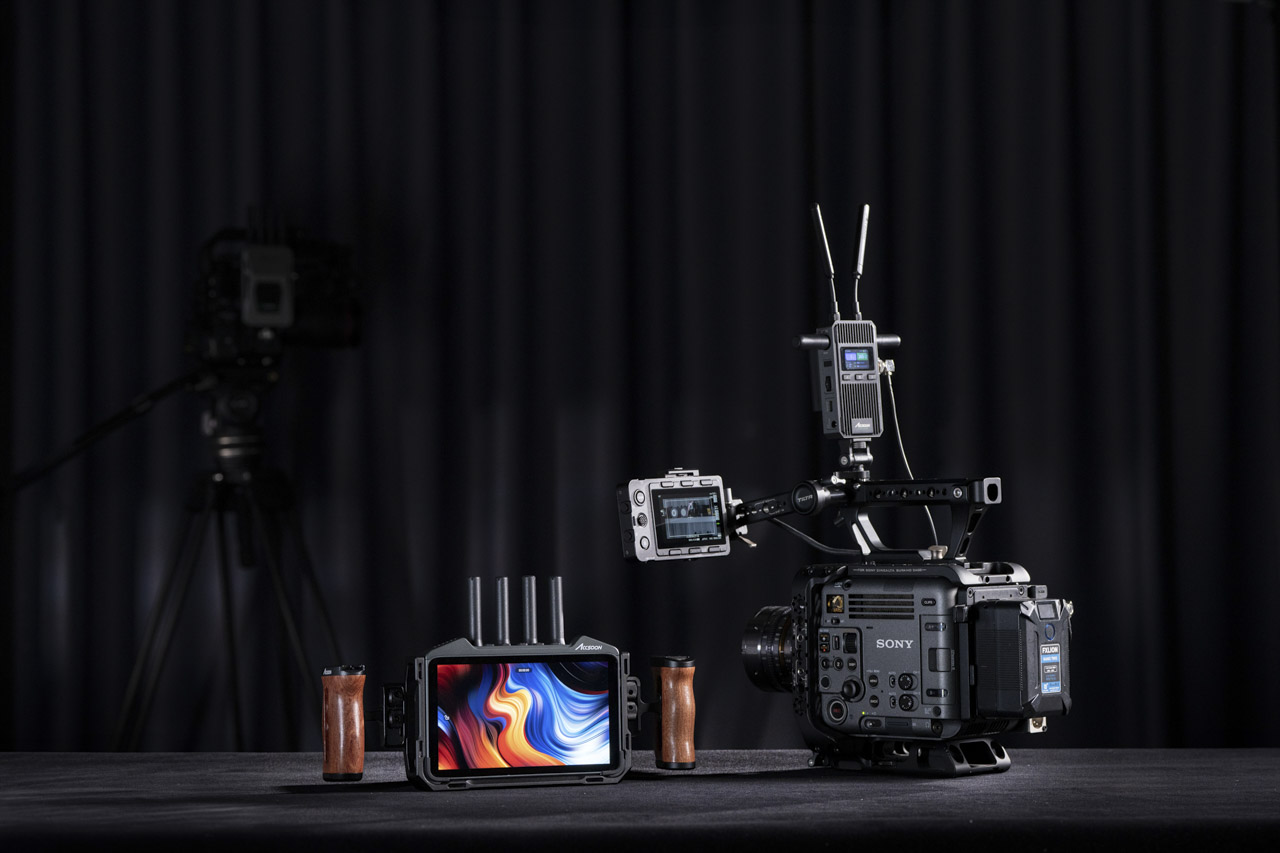 A monitor with blue and orange waves on the screen and prongs on the top sitting next to a professional cameras with attachments affixed