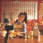 A girl wearing a yellow sweater sits in front of a camera holding a fork and eating