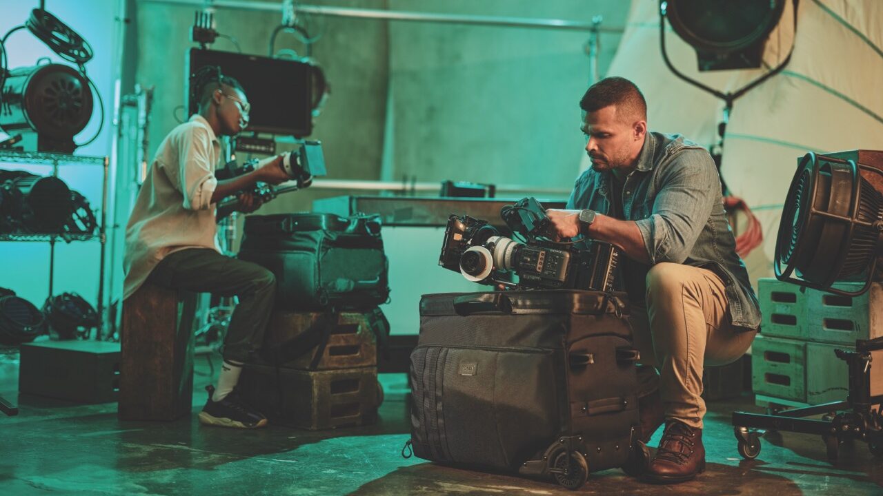 Two people sitting next to kit bags in a green room, looking at cameras