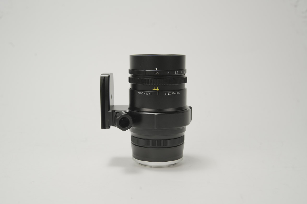 A black macro lens from Mitakon sitting vertically on a table with a handle on the left