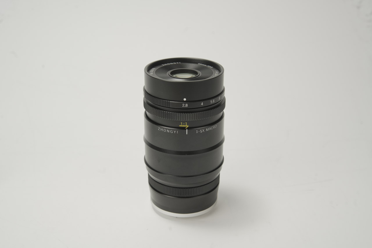 A black macro lens from Mitakon sitting vertically on a white surface