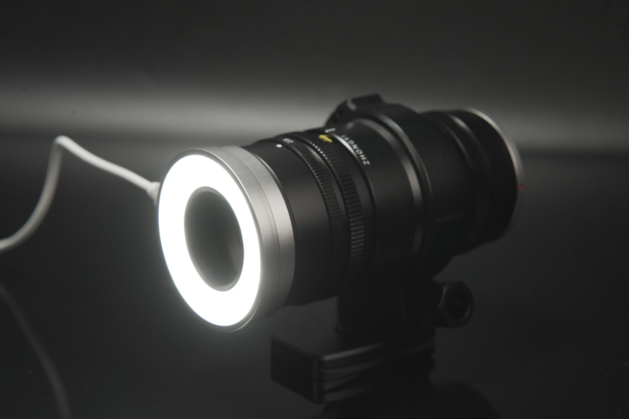 A black macro lens from Mitakon with a glowing rim and a wire attached, against a dark background