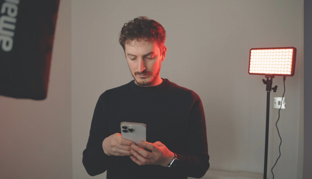 A man standing in a studio with a rectangular amaran light behind him, looking at his phone
