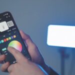 A hand adjusts a colour wheel on a smartphone. A rectangular light from amaran stands in the background